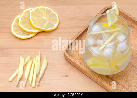 several slices of lemon, light-brown ginger cut in pieces and a water glass full of cold refreshing lemonade with ice cubes on a wooden tray, healthy energy drink ready be served with ingredients Stock Photo