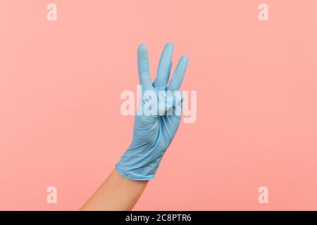 Profile side view closeup of human hand in blue surgical gloves showing number 3 three with hands. indoor, studio shot, isolated on pink background.