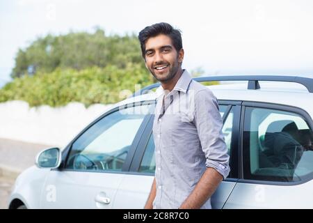 Happy man driver smiling standing by his new sport blue car isolated outside parking lot background. Handsome young man excited about his new vehicle. Stock Photo
