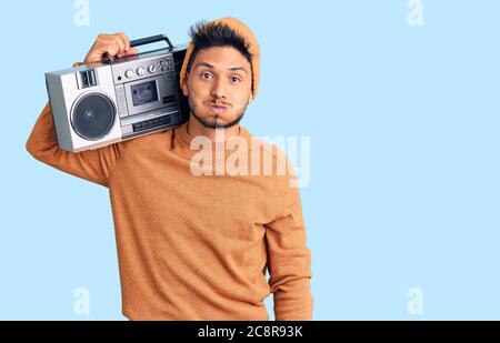 Handsome latin american young man holding boombox, listening to music puffing cheeks with funny face. mouth inflated with air, crazy expression. Stock Photo