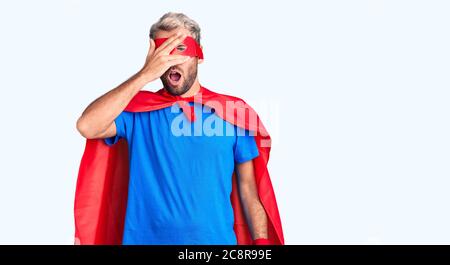 Young blond man wearing super hero custome peeking in shock covering face and eyes with hand, looking through fingers with embarrassed expression. Stock Photo