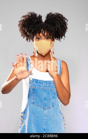 Afro-Brazilian teenage model gestures with a stop hand and points to the use of a protective mask against invite-19 Stock Photo