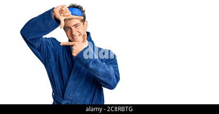 Young handsome man wearing sleep mask and bathrobe smiling making frame with hands and fingers with happy face. creativity and photography concept. Stock Photo