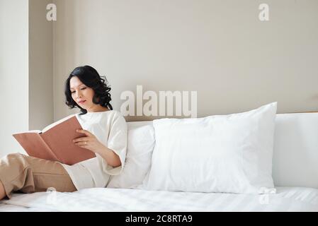 Pretty Asian woman sitting on bed and reading interesting book Stock Photo