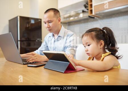 Serious Vietnamese man answering e-mails on laptop when daughter sitting next to him and using educational application on tablet computer