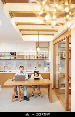 Serious Vietnamese man working on laptop at kitchen table when daughter sitting next to him and doing homework or having online class due to lockdown Stock Photo