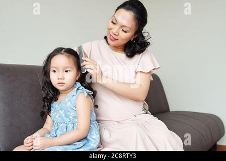 Pensive little girl sitting on sofa when mother combing her curled hair Stock Photo