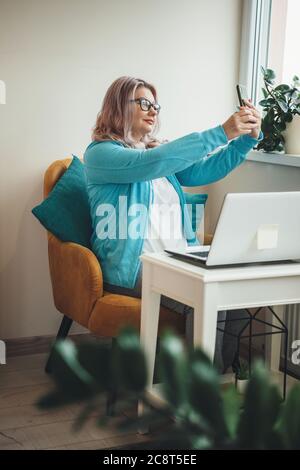 Senior caucasian businesswoman with eyeglasses making a selfie sitting in armchair and using a laptop Stock Photo