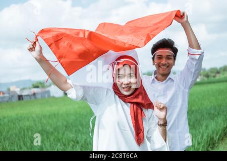 a boy and hijab girl wearing red and white attributes raised the Indonesian flag when standing outside in the rice fields Stock Photo