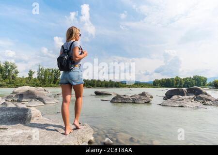 Blonde in beautiful denim shorts with a black backpack standing by the river on the rocks. Portrait from the back of the girl traveler. Photo travel Stock Photo