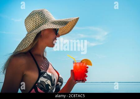 A young woman in sunglasses and a straw hat holds a cocktail in her hand against the blue sky and sea. Beautiful girl is drinking ice summer drink in