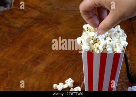 Hand taking popcorn out of red and white striped cardboard bucket retro design on wooden table Stock Photo