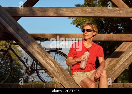 A middle-aged woman sits on a wooden bridge, rests after cycling and drinks water. Stock Photo