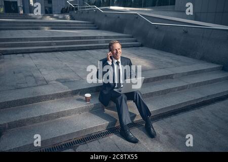 Joyful man using laptop and talking on cellphone outdoors Stock Photo