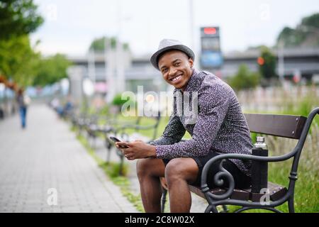 Cheerful young black man commuter sitting on bench outdoors in city. Stock Photo