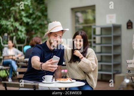 Happy young couple sitting outdoors in cafe, taking selfie by smartphone. Stock Photo