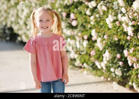 Funny child girl 2-3 year old smiling over rose flower background closeup. Looking at camera. Childhood. Summer season. Happiness. Stock Photo