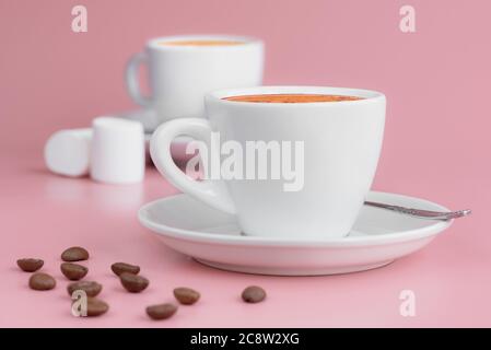 Two white cups of hot aroma coffee, some marshmallows and coffee beans on pink background. Morning coffee concept Stock Photo