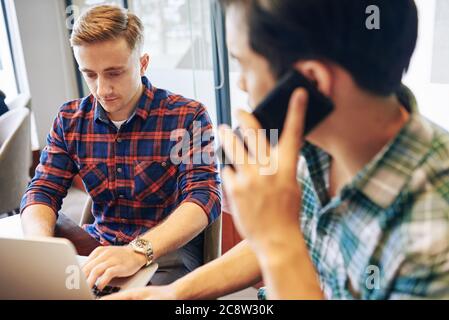 Serious concentrated young man programming on laptop when his coworker talking on phone Stock Photo