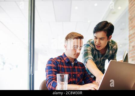 Handsome young Vietnamese man pointing at mistake of colleague in programming code on laptop screen Stock Photo