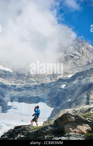 Side view of traveler in shorts standing on rocky hill and holding professional camera. Man hiker admiring the view of beautiful snowy mountains. Concept of travelling, hiking and mountaineering. Stock Photo