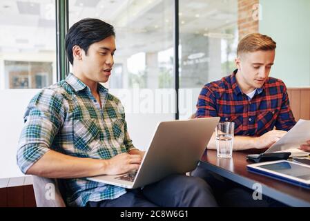 Vietnamese young man checking e-mail on laptop when his coworker reading report Stock Photo