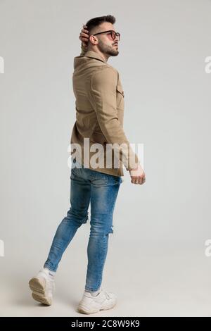 back view of young casual guy wearing glasses, looking to side and scratching head, thinking, walking on grey background Stock Photo