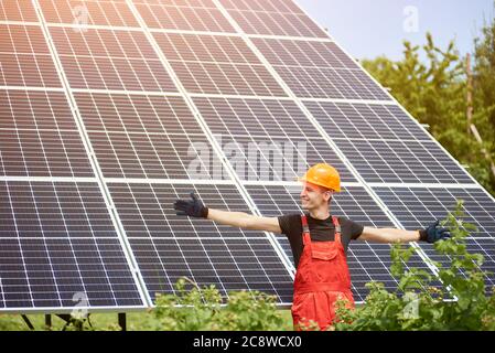 Young man smiling with arms open against the background of solar panels, he satisfied his work. A male is wearing an orange uniform, hard hat, black t-shirt and gloves on a warm day Stock Photo