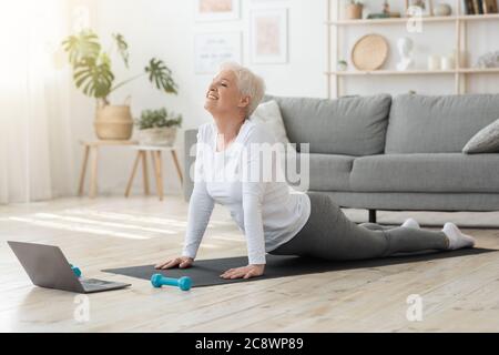 Online Yoga. Senior Woman Standing In Cobra Pose In Front Of Laptop Stock Photo