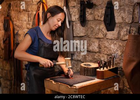 Leather craftswoman in her atmospheric workshop working on a hand-made belt. concept: female artisan lifestyle Stock Photo