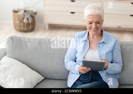 Charming elderly lady sitting on couch at home using digital tablet computer Stock Photo