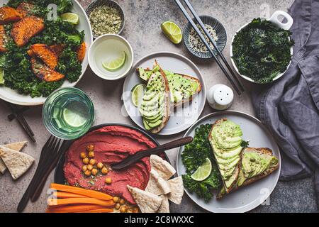 Vegan lunch table, top view. Flat-lay of baked sweet potato salad, avocado toasts and hummus on dark background. Stock Photo