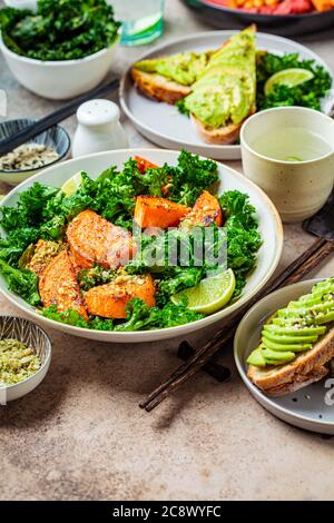 Vegan lunch table. Baked sweet potato salad with kale, avocado toast and hummus on a dark background. Stock Photo