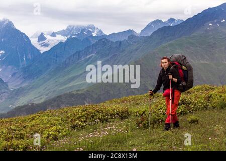 Girl hiker with hiking backpack in mountains. Tourist in Svaneti on a background of the main Caucasus ridge and snow-capped mountain peaks. Stock Photo