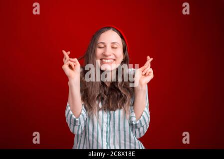Cheerful young woman is crossing her fingers like she is hoping for something over red background. Stock Photo