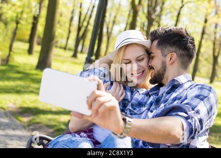 Happy caucasian couple sitting on a park bench, taking cute selfies during daylight Stock Photo