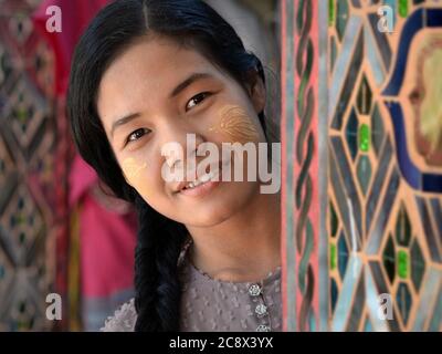 Pretty Burmese girl with natural thanaka face cosmetic on her cheeks peeks from behind a pillar at the Buddhist Su Taung Pagoda. Stock Photo