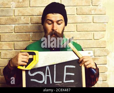 bearded worker man, long beard, brutal caucasian hipster with moustache holding various building tools: saw, hammer and board with inscription sale, surprised face, brick wall studio background Stock Photo