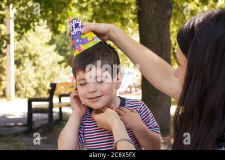 A mother puts a festive cap on her son's head to a child of 3 years. Birthday, children's holiday in the park Stock Photo