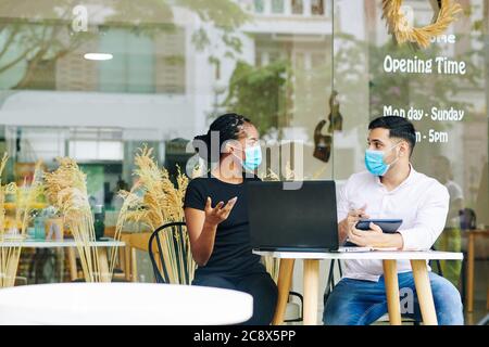 Multi-ethnic entrepreneurs in medical masks meeting at cafe table and discussing business strategy Stock Photo