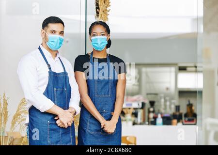 Young waiter and waitress in medical masks and aprons standing at entrance of small bakery Stock Photo