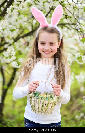 Girl smiling in bunny ears with basket. Child playing in garden with blossoming trees. Childhood, youth and growth. Happy easter and spring concept. Holidays celebration with presents and eggs. Stock Photo