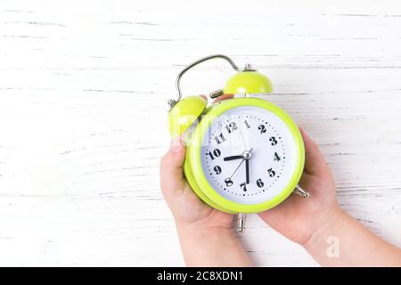 Kid hands holding green vintage alarm clock, top view, white wooden background with copy space Stock Photo