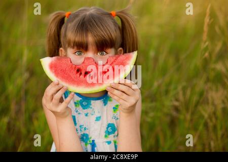 Happy child with big red slice of watermelon sitting on green grass in summer park. Healthy eating concept Stock Photo