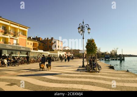 View of the lakeside promenade with people sitting in outdoor cafe and walking with dogs in a sunny winter day, Lazise, Verona, Veneto, Italy Stock Photo