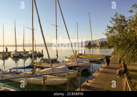 View of the port on the lake shore with sailboats and people on benches and walking on the lakeside promenade at sunset, Bardolino, Veneto, Italy Stock Photo