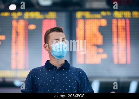 Man wearing face mask against airport departure board. Themes new normal, coronavirus and personal protection. Stock Photo