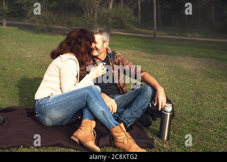 Happy middle-aged couple in love sitting on a blanket in the grass in park, hug, look at each other, smile and drink tea or coffee from thermos. Stock Photo
