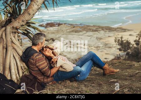 Happy middle-aged couple in love sitting under the tree with ocen view, hug, smile and look at each other. Stock Photo