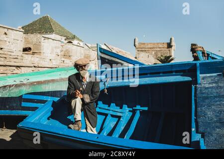 ESSAOUIRA, MOROCCO - SEPTEMBER 10, 2019: Old senior fisherman on blue wooden boat in Essaouira harbor. Stylish look, hat, beard, sunglasses and jacket. Moroccan people. Stock Photo